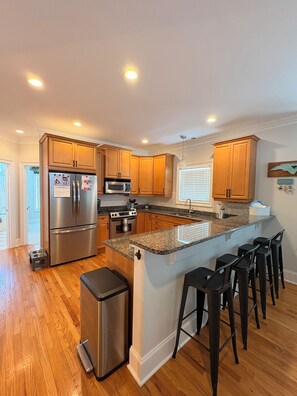 A modern kitchen with wooden cabinets, stainless steel appliances, a granite countertop, and a bar area with stools. - 6 BR Townhome in Carolina Beach with Public Beach Access across the Street! (Carolina Beach)
