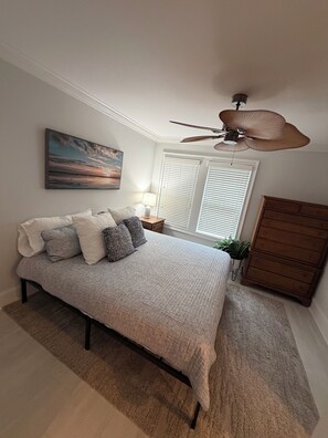Une chambre à coucher avec un lit, un ventilateur de plafond, une fenêtre avec des stores, une commode et un tableau au mur.