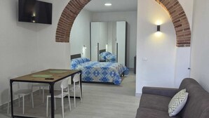 Ein modernes Wohnzimmer mit Sofa, einem Esstisch mit Stühlen, einem an der Wand befestigten Fernseher sowie ein Schlafzimmer mit Bett und Spiegel.
