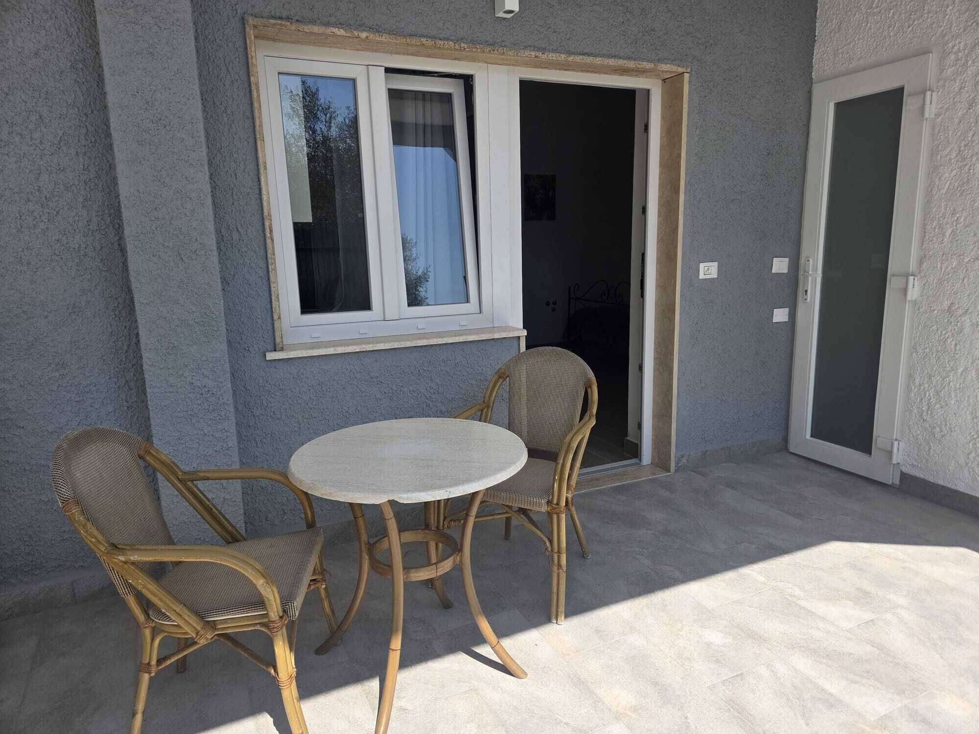 An outdoor patio with a table and two wicker chairs, a window with a curtain, and a door leading to an interior space.