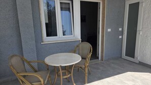 Eine Terrasse im Freien mit Tisch und zwei Korbstühlen, ein Fenster mit Vorhang und eine Tür, die in einen Innenraum führt.