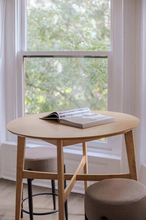 A small wooden table with a book and a notebook, a cushioned stool, and a window with a view of trees. - Designer Loft 3E | Heart of Gold Coast/River North (Chicago)