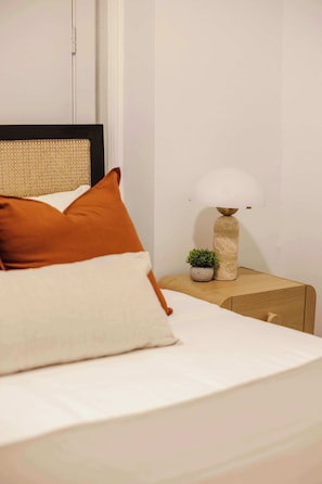 A neatly made bed with white bedding and two orange pillows, a small potted plant, and a lamp on a wooden nightstand. - Designer Loft 4E | Heart of Gold Coast/River North (Chicago)