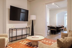 Un salon moderne avec un téléviseur à écran plat fixé au mur, une table basse ronde sur laquelle repose un livre, une console noire agrémentée d’objets décoratifs et, au fond, un coin repas.