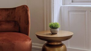 A room with a single brown chair, a small round table with a plant, and a window with a view of buildings. - Designer Loft 4E | Heart of Gold Coast/River North (Chicago)
