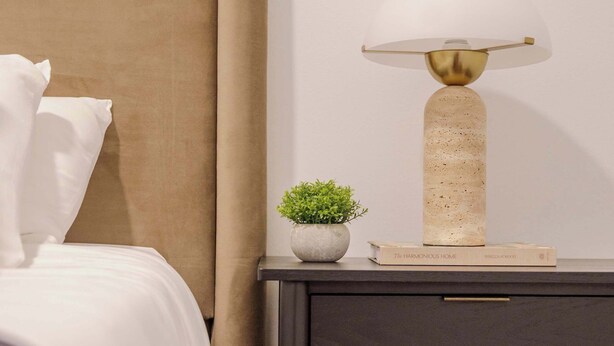 A neatly made bed with white linens, a beige headboard, and a small bedside table with a lamp, a potted plant, and a book titled 'The Harmonious Home'. - Designer Loft 4E | Heart of Gold Coast/River North (Chicago)
