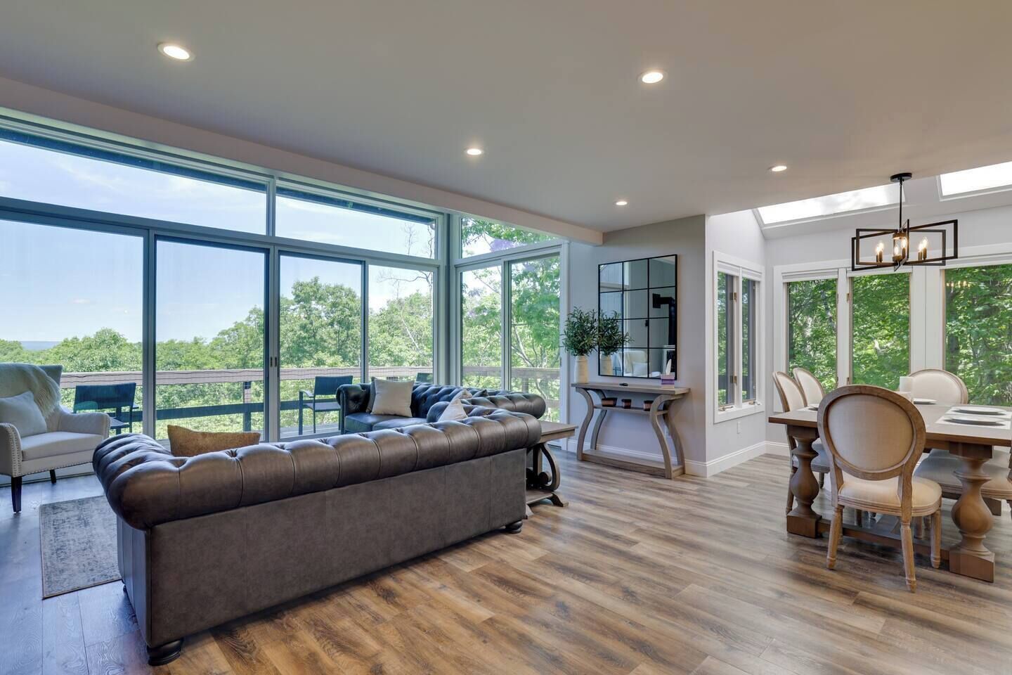 A spacious living room with a large gray sofa, a dining area with a wooden table and chairs, and large windows offering a view of the outdoors.