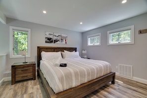 A bedroom with a large bed, wooden headboard, and a nightstand with a lamp. There are two windows with white frames and a wall-mounted heater. - Luxury Poconos Townhouse with Resort Perks! (Tannersville)
