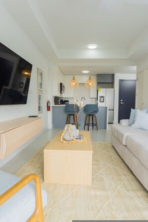 A modern living room with a flat-screen TV, a wooden coffee table, a sofa, and a kitchen in the background. - Solèa 6th-Floor High-Rise Studio | Panoramic Ocean Views & Walk to Town (Ocho Rios)