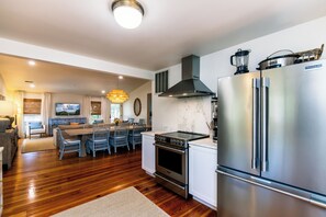 A modern kitchen with a stainless steel refrigerator, a built-in oven, and a stove. There is a dining area with blue chairs and a chandelier. - Gas Money | Private Pool + Spa | Screened Porch & Sun Decks | 5-Minute Beach Walk | Sleeps 9 (Edisto Beach)