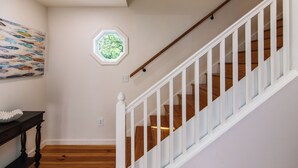 Una escalera con barandillas blancas y peldaños de madera, una pared con un ventanal hexagonal, y un cuadro de peces en la pared.