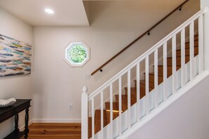 Un escalier avec des rampes blanches et des marches en bois, un mur orné d’une fenêtre hexagonale et un tableau représentant des poissons.