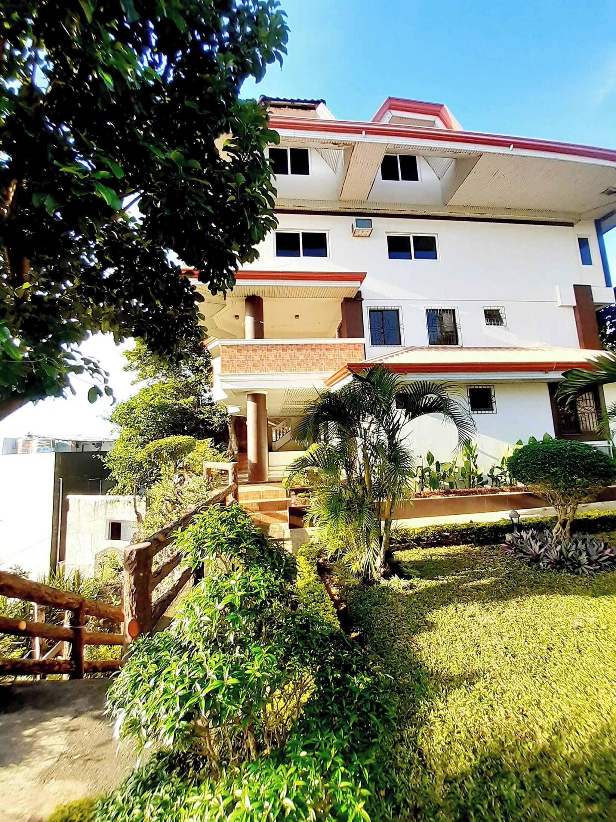 A multi-story building with a red roof, surrounded by lush greenery and a wooden fence.