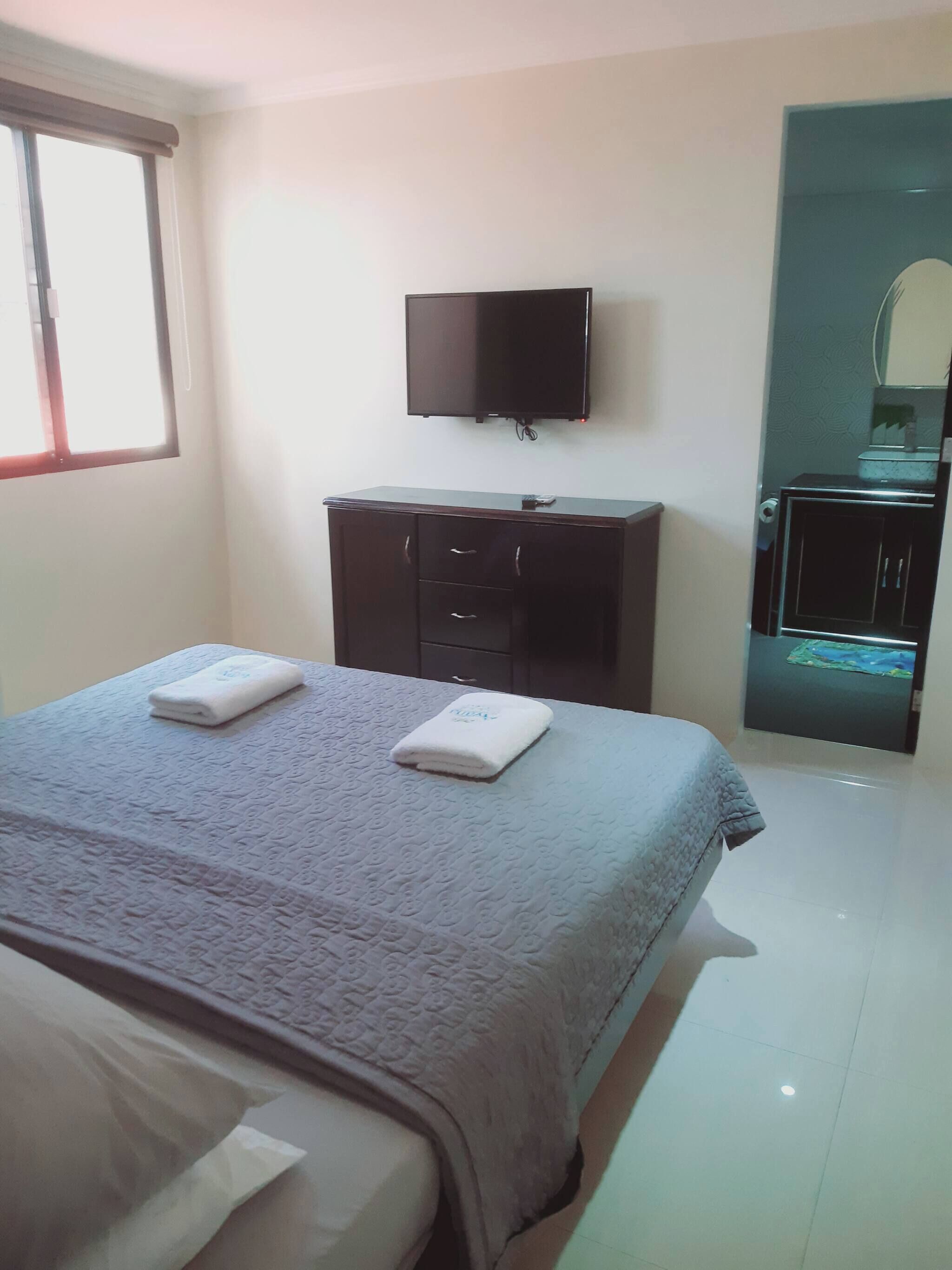 Habitación de hotel con una cama, un televisor de pantalla plana montado en la pared, un aparador de madera oscura y un baño con espejo y lavabo.
