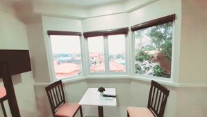 Una camera d'angolo con un tavolo e due sedie, ampie finestre che si affacciano su edifici e alberi, e una lampada a soffitto.
