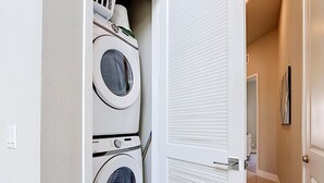 Uma lavanderia com duas máquinas de lavar empilhadas, uma porta que dá para um corredor e um porta-toalhas fixado na parede.