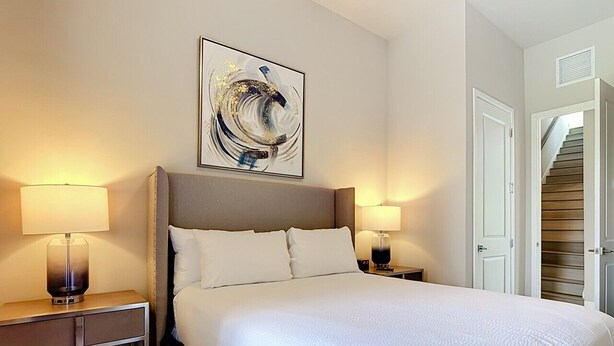 Quarto de hotel com uma cama grande, duas mesas de cabeceira com abajures, um quadro abstrato na parede e uma porta que dá para uma escada.