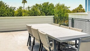 Um pátio externo com mesa e cadeiras, cercado por um muro baixo e vegetação.