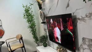 Ein modernes Wohnzimmer mit einem Flachbildfernseher an einer Marmorwand, einer weißen Unterhaltungseinheit, einem Holzboden, einem schwarzen Metallstuhl und einer Topfpflanze.