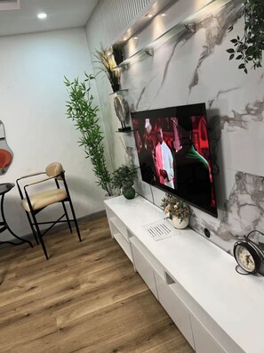 A modern living room with a flat-screen TV mounted on a marble wall, a white entertainment unit, a wooden floor, a black metal chair, and a potted plant. - Radiant 1-Bedroom Apartment in Lekki 1 with Fast Wi-Fi & 24/7 Power (Lagos)