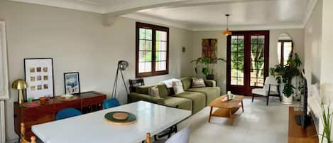 Un vaste espace de vie comprenant une table à manger, des chaises, un canapé et une table basse. Une grande fenêtre offre une belle lumière naturelle et une vue sur la verdure environnante.