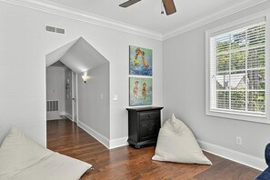 Een kamer met houten vloeren, een plafondventilator, een raam met jaloezieën en kunst aan de muur.