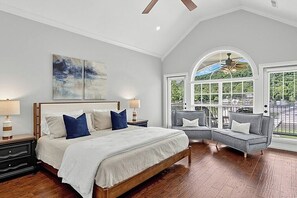 A bedroom with a large bed, two bedside tables, a gray sofa, and a window with a view of greenery. - Waters Edge- Lakefront Luxury on Lake Guntersville (Grant)