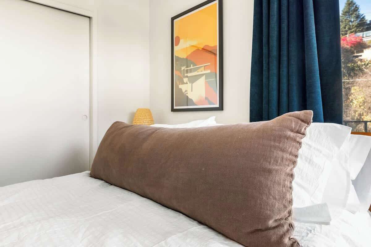 Ein Bett mit weißer Bettdecke und braunem Kopfkissen, ein gerahmtes Bild an der Wand und ein Fenster mit Vorhängen.