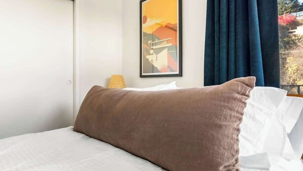 Una cama con sábana blanca y almohada marrón, un cuadro en la pared y una ventana con cortinas.