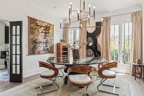 A dining area with a glass table surrounded by modern chairs, a chandelier, and a large abstract painting on the wall. - The Black Fig | Hollywood Character Modern Comfort (Los Angeles)