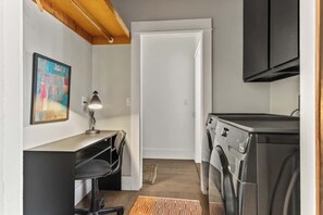 A compact laundry room with a washing machine, a desk with a chair, a lamp, and a door leading to another room. - The Black Fig | Hollywood Character Modern Comfort (Los Angeles)