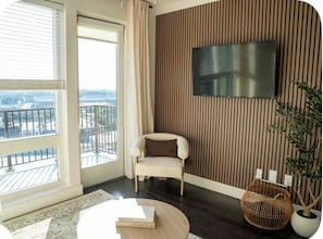 Una sala de estar moderna con un ventanal grande, un televisor de pantalla plana montado en una pared de madera, un sillón cómodo, una mesa de centro redonda y una mesita auxiliar de mimbre con una planta en maceta.