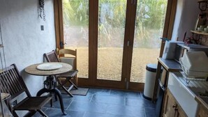 A small kitchen with a round table and chairs, a trash bin, and a view of a garden through the door.