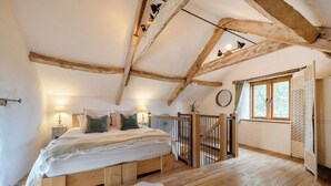Ein Schlafzimmer mit Holzbalkendecke, einem Bett mit Kissen, einem Nachttisch, einem Fenster mit Vorhängen und Holzboden.