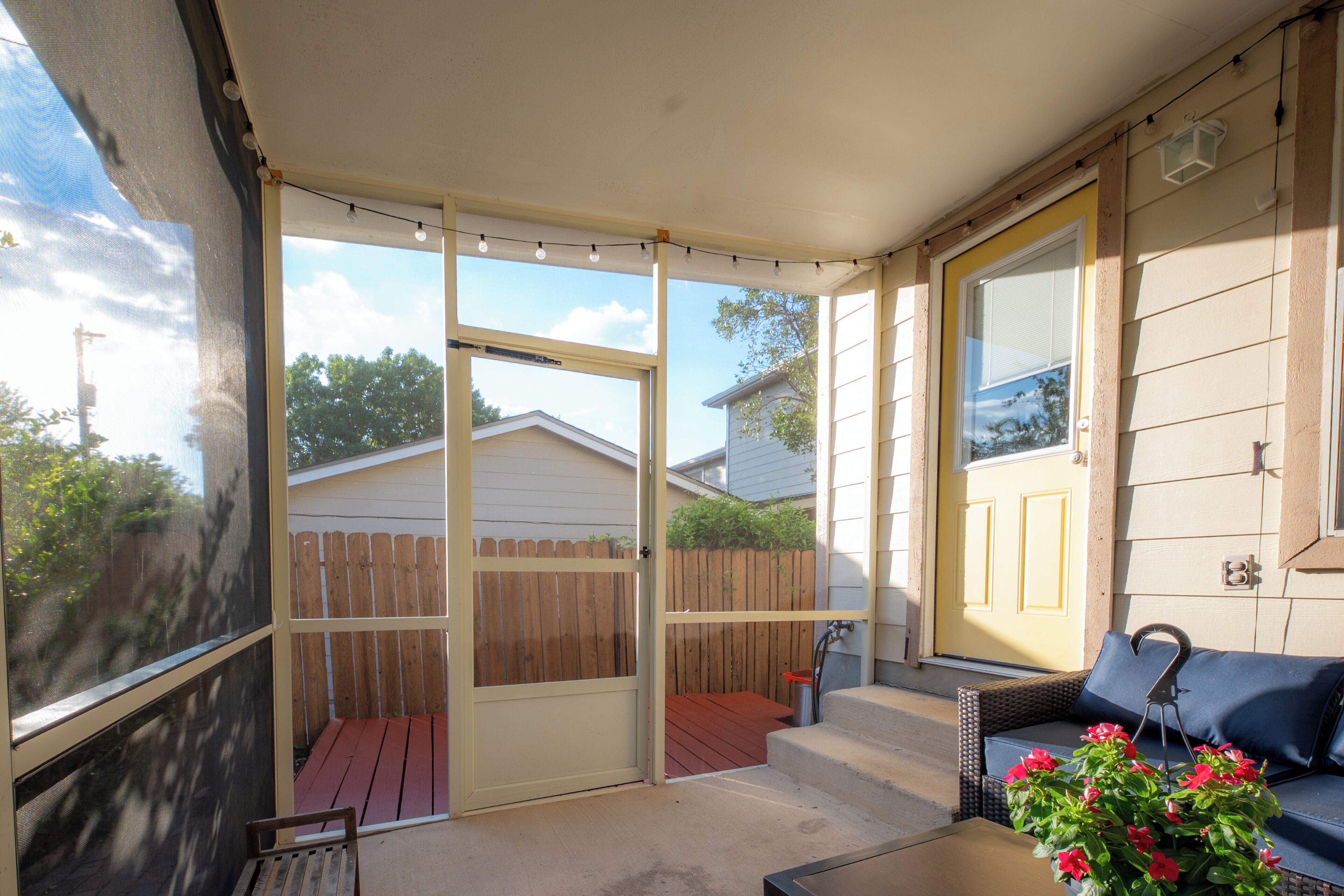 Une terrasse couverte avec une porte-fenêtre coulissante, une terrasse en bois, une table rouge et un canapé en osier agrémenté d’un arrangement floral.