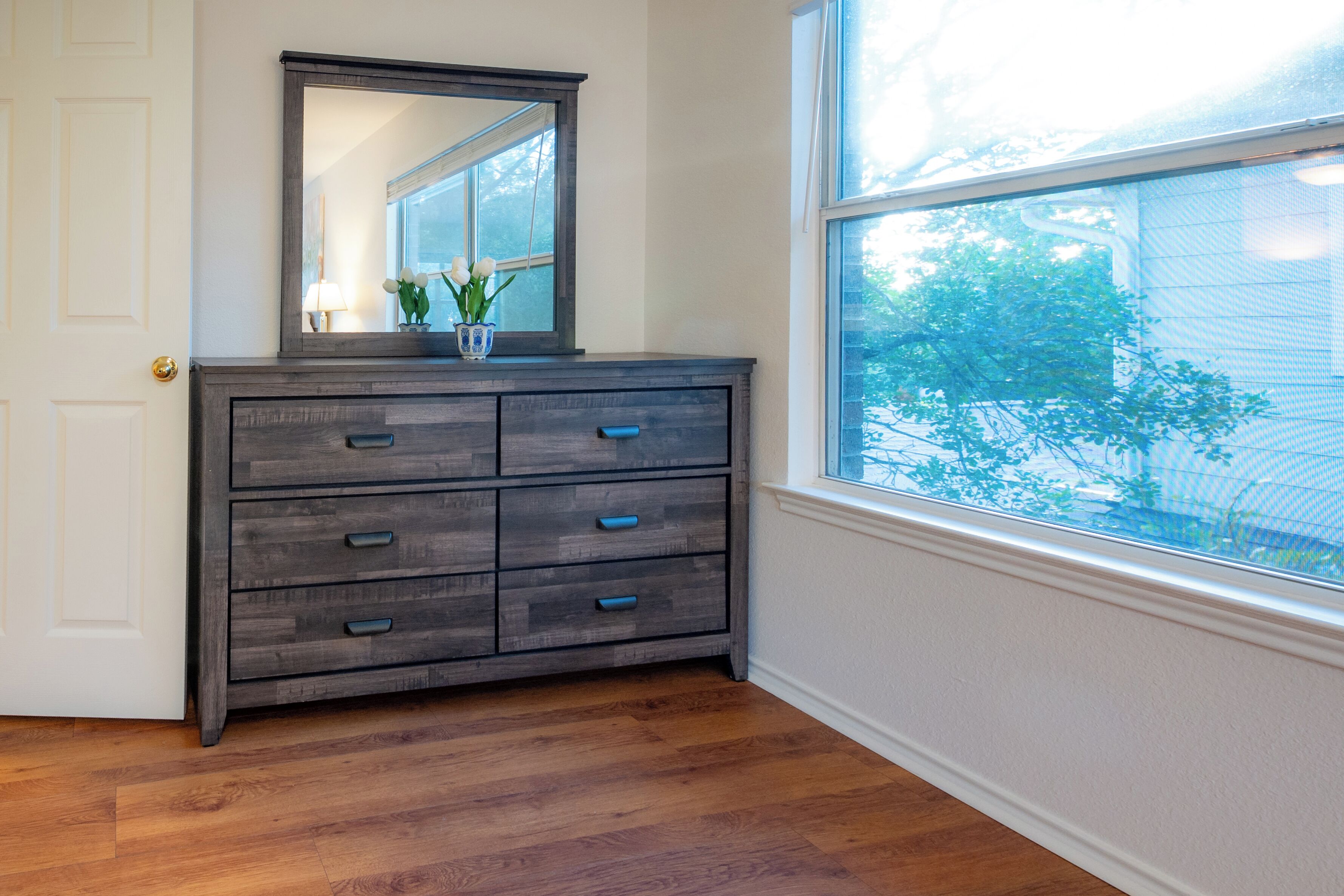 A dark wooden dresser with multiple drawers against a white wall, positioned next to a window with a view of trees.