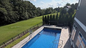 Ein rechteckiger Pool mit einer Steindeckplatte, umgeben von einem schwarzen Metallzaun und einem Rasenbereich mit Bäumen im Hintergrund.