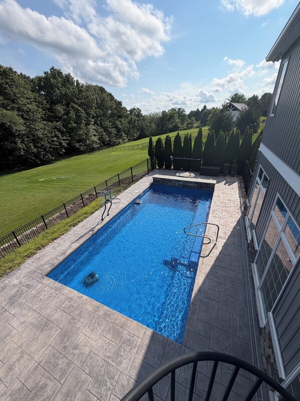 En rektangulär swimmingpool med en stenbelagd poolkant, omgiven av ett svart metallstaket, och en gräsyta med träd i bakgrunden.