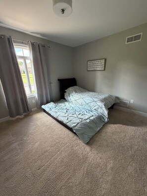 A bedroom with a bed, curtains, a wall-mounted air vent, and a ceiling light fixture.