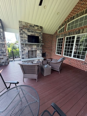 A covered outdoor area with a stone fireplace, wicker furniture, and large windows. - Tranquil luxurious 4-bedroom home on a scenic 2.5 ace lot, min. from Penn State. (Port Matilda)