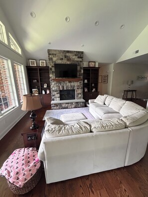 A living room with a stone fireplace, a large white sofa, and a television mounted above it. - Tranquil luxurious 4-bedroom home on a scenic 2.5 ace lot, min. from Penn State. (Port Matilda)