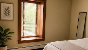 A bedroom with a wooden bed, a window with blinds, a potted plant, a framed picture, and a mirror.