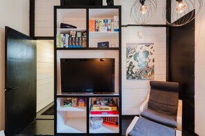 A living room with a flat-screen TV, shelves with board games, a chair, and a wall art piece. - Cozy Sunriver Retreat - SHARC Passes, Bike and BBQ (Sunriver)