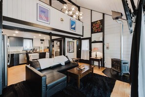 A modern living room with a black leather sofa, a wooden coffee table, and a black rug. There is a kitchen visible in the background with white cabinets and a dining area. - Cozy Sunriver Retreat - SHARC Passes, Bike and BBQ (Sunriver)