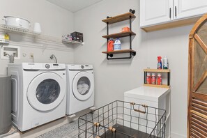 A laundry room with two white Samsung washing machines, a small sink, and shelves with cleaning supplies.