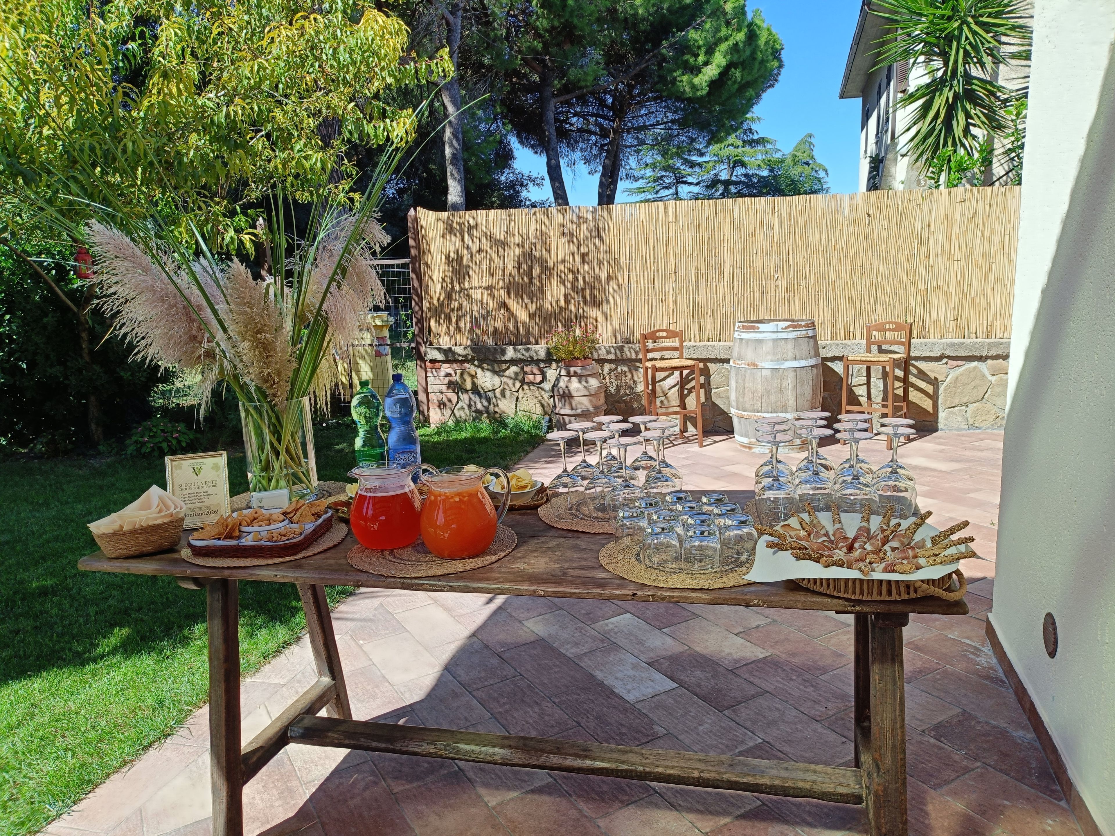 Eine Terrasse im Freien mit einem Holztisch, gedeckt für eine Mahlzeit, mit Brot, Gebäck und Getränken.