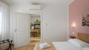 Habitación de hotel con cama, escritorio, silla, una mesita, un cuadro y una cocina visible a través de una puerta abierta.