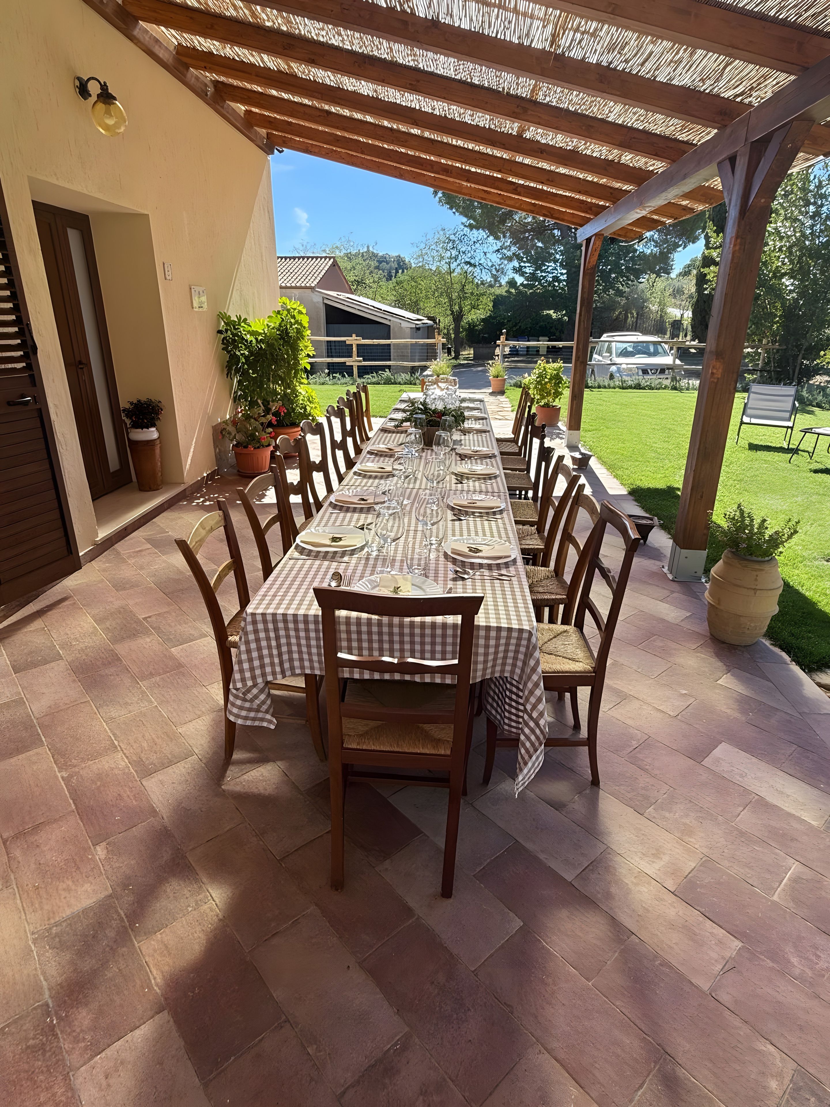 Une terrasse extérieure avec une longue table à manger, dressée pour un repas, entourée de chaises et abritée par une pergola en bois.