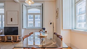 A modern living room with a glass dining table, leather chairs, a flat-screen TV, and a large window with a view of buildings. - Downtown 2BR in Santa Justa by LovelyStay (Lisboa)