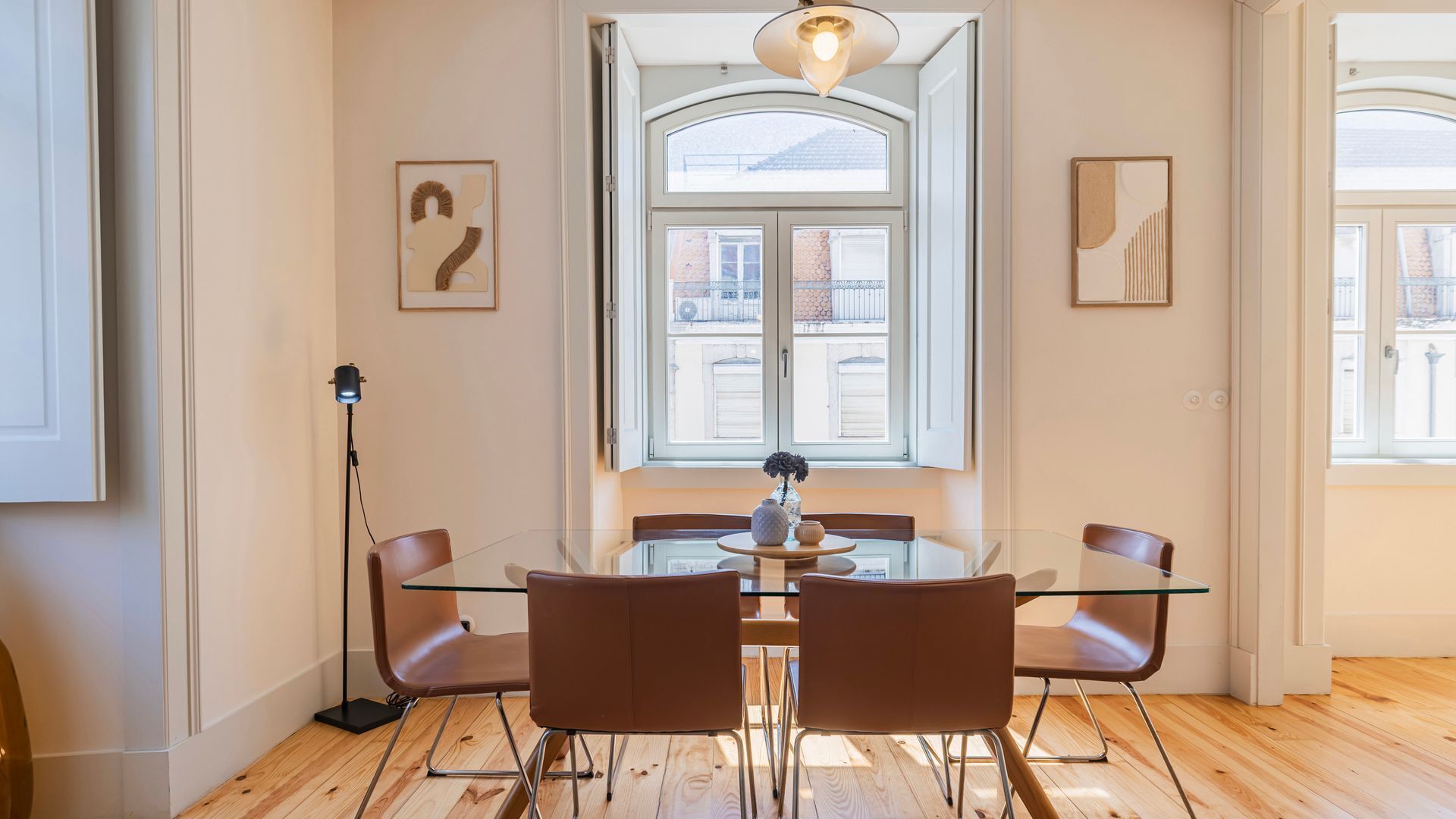 A modern dining area with a glass table and brown chairs, a floor lamp, and a large window with a view of buildings.